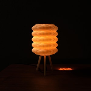 Puede incluir: Una lámpara crema acanalada con una pantalla cilíndrica, iluminada desde dentro, apoyada sobre una base de madera de tres patas. La lámpara proyecta un brillo naranja cálido sobre un fondo oscuro, creando un ambiente acogedor.