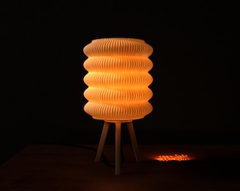 May include: A cream-colored, ribbed lamp with a cylindrical shade, illuminated from within, resting on a three-legged wooden base. The lamp casts a warm, orange glow against a dark background, creating a cozy ambiance.