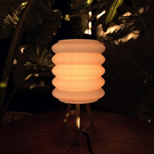 K&ouml;nnte beinhalten: Eine cremefarbene, gerippte Lampe mit einem gestapelten, abgerundeten Design. Die Lampe steht auf einem dreibeinigen Holzst&auml;nder. Die Lampe ist beleuchtet und wirft einen warmen Schein. Der Hintergrund zeigt dunkelgr&uuml;nes Laub.