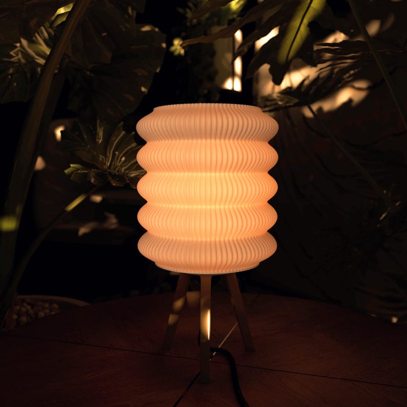 K&ouml;nnte beinhalten: Eine cremefarbene, gerippte Lampe mit einem gestapelten, abgerundeten Design. Die Lampe steht auf einem dreibeinigen Holzst&auml;nder. Die Lampe ist beleuchtet und wirft einen warmen Schein. Der Hintergrund zeigt dunkelgr&uuml;nes Laub.