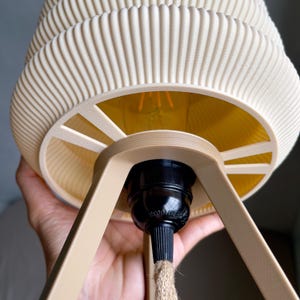 K&ouml;nnte beinhalten: Eine beige, gerippte Pendelleuchte mit warmgelbem Innenraum. Die Leuchte hat eine schwarze Fassung und einen beigen Rahmen. Ein Seil h&auml;ngt aus der Fassung.