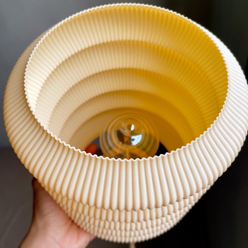 K&ouml;nnte beinhalten: Cremefarbener, geschichteter Lampenschirm mit gerilltem, spiralf&ouml;rmigem Design. Die Lampe hat einen warmen, gelben Schein von der Gl&uuml;hbirne im Inneren. Der Schirm wird von einer Hand gehalten und zeigt seine einzigartige, strukturierte &Auml;sthetik. Eine moderne Leuchte.