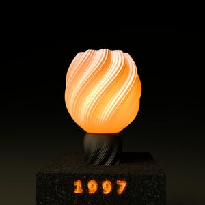 Puede incluir: Lámpara esférica con un diseño en espiral y en capas, en naranja y blanco. La lámpara está sobre una base negra texturizada con el año "1997" iluminado en naranja. El diseño es moderno y artístico, con un brillo cálido.