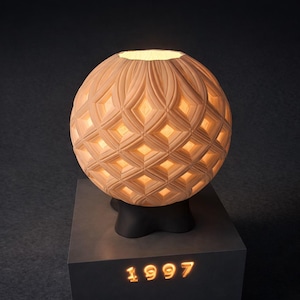 May include: A beige spherical lamp with a diamond pattern, radiating a warm light. The lamp is placed on a dark grey pedestal, with the illuminated year "1997" at the base. The lamp's design features a carved, openwork structure.