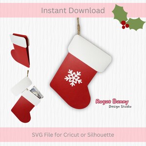 Puede incluir: Calcetín de Navidad rojo y blanco con un diseño de copo de nieve. El calcetín está colgado de una cuerda y está abierto para revelar un interior blanco. El texto "Rogue Bunny Design Studio" es visible en la esquina inferior derecha de la imagen. Archivo SVG para Cricut o Silhouette.