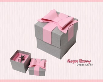 May include: A gray gift box with a pink bow on top. The box is open and there is a small silver figurine inside. The box is labeled "Rogue Bunny Design Studio" and "SVG File for Cricut or Silhouette".