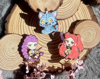 May include: Three enamel pins featuring cartoon characters. One is a blue cat-like creature with yellow eyes, and the other two are female characters with purple and red hair, holding weapons. The pins are displayed on wooden slices.