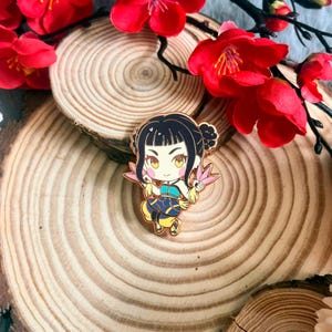 Puede incluir: Un pin de esmalte dorado que representa un personaje de dibujos animados con cabello negro, ojos amarillos y un atuendo azul y amarillo. El pin está decorado con accesorios rosas en forma de abanico. El pin está sobre una superficie de madera con flores rojas.