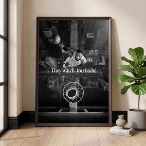 Könnte beinhalten: Schwarz-Weiß-Druck mit Rahmen, der einen Basketballspieler im Lakers-Trikot vor dem Hintergrund einer Stadt zeigt. Der Text "They watch. You build." ist zu sehen. Ein Basketballkorb befindet sich im Vordergrund.