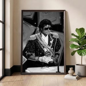 May include: Black and white framed print of Michael Jackson in a formal jacket with epaulettes, sunglasses, and a sequined glove, holding an award. The image is displayed in a room with a window and a potted plant.