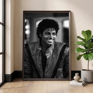 Op de afbeelding: Zwart-wit ingelijst portret van Michael Jackson die lacht. Hij draagt een leren jas. Het portret staat op een houten vloer, naast een plant in een pot en decoratieve items.