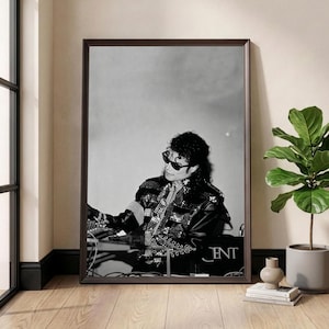May include: Black and white framed print featuring a portrait of a musician wearing sunglasses and a studded jacket. The print is displayed in a room with a window and a potted plant. The print has the word "TNT" on it.