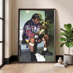 May include: Framed print featuring a seated figure in a purple and gold jacket, holding a gold trophy. The image is set in a locker room with a tiled wall. The print is displayed in a dark brown frame, adding a touch of elegance to the sports-themed artwork.