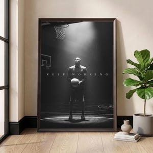 May include: Black and white framed print featuring a basketball player holding a ball on a court. The words "KEEP WORKING" are displayed above the player. The image is set in a dark, dramatic setting with a spotlight.