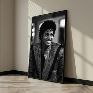May include: Black and white framed portrait of a smiling man with curly hair. He is wearing a leather jacket with a high collar. The portrait is leaning against a wall.