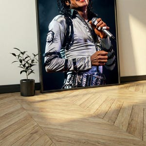 May include: Framed print of a person wearing a silver jacket with black accents and a microphone in hand. The person is smiling. The print is in a black frame and leans against a white wall. A small potted plant sits to the left.