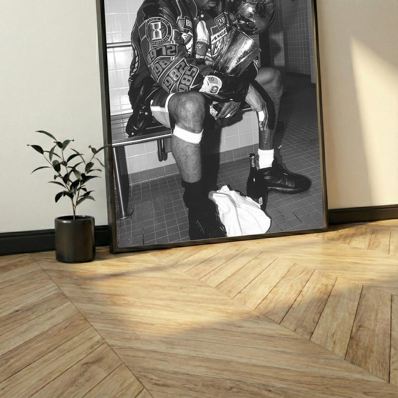 May include: Black and white framed print featuring a person in a jacket holding a trophy. The print is displayed on a wall, with a small potted plant in the foreground. The image is in a black frame.