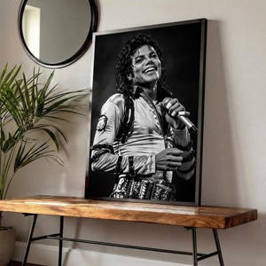 May include: Black and white framed print of Michael Jackson smiling and holding a microphone. He is wearing a light-colored shirt with a decorative belt. The print is displayed on a wooden bench with black metal legs.