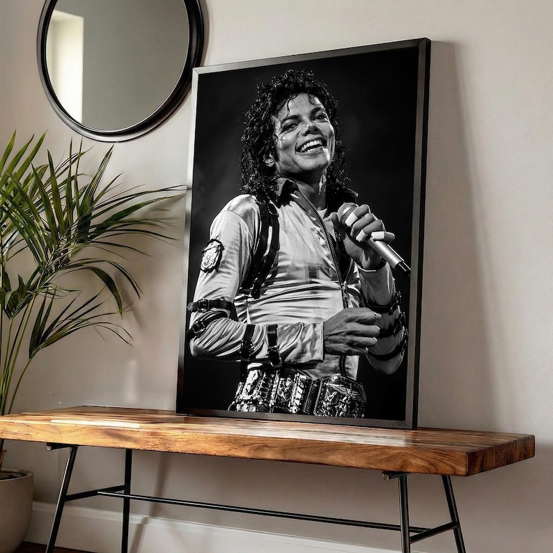 May include: Black and white framed print of Michael Jackson smiling and holding a microphone. He is wearing a light-colored shirt with a decorative belt. The print is displayed on a wooden bench with black metal legs.