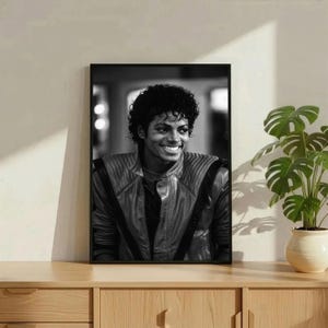 May include: Black and white framed portrait of a smiling man with curly hair. He is wearing a leather jacket with a quilted design. The portrait is displayed on a wooden cabinet, with a potted plant to the right.