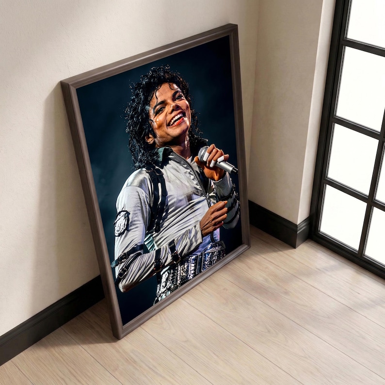May include: Framed portrait of Michael Jackson in a silver jacket, smiling and holding a microphone. The image is in a brown frame, leaning against a white wall and wooden floor.