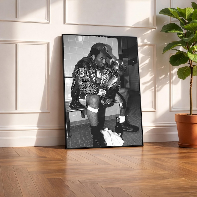 May include: Black and white framed photograph of a man in a leather jacket and baseball cap, holding a trophy. The man is seated on a bench, with a towel and bottle nearby. The image is leaning against a white wall.
