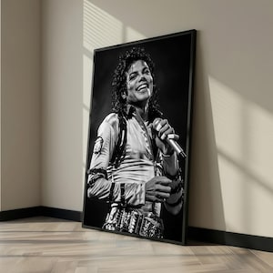 May include: Black and white framed print of a smiling person holding a microphone. The person is wearing a light-colored shirt with a detailed belt and wrist cuffs. The image is leaning against a wall.