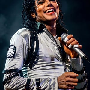 May include: A person wearing a silver jacket with black straps and a decorative belt, holding a microphone. The jacket has a zipper and a patch on the left sleeve. The person is smiling.