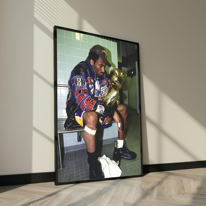 May include: Framed print featuring a seated figure in a jacket, holding a golden trophy. The jacket is purple and gold with patches. The figure is wearing a baseball cap and shorts. The background is a tiled wall.