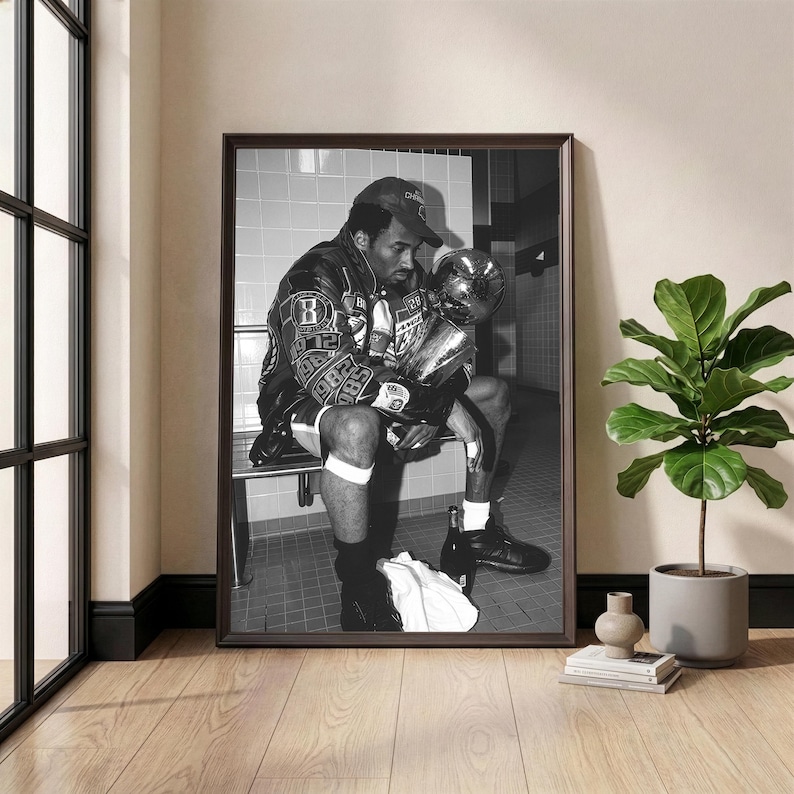 May include: Black and white framed photograph of a person seated on a bench, holding a trophy. The person is wearing a jacket with patches and a baseball cap. The photograph is displayed in a room with a window and a potted plant.