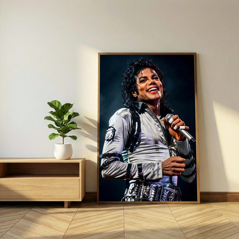 May include: A framed portrait of Michael Jackson, smiling and holding a microphone. He wears a silver jacket with black accents and a decorative belt. The portrait is displayed on a wall next to a wooden cabinet and a potted plant.