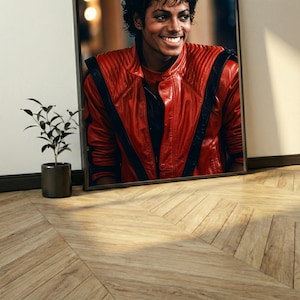 May include: Framed portrait of Michael Jackson smiling, wearing a red leather jacket with black stripes. The portrait is leaning against a white wall, with a small potted plant to the left. The floor is made of wooden planks.