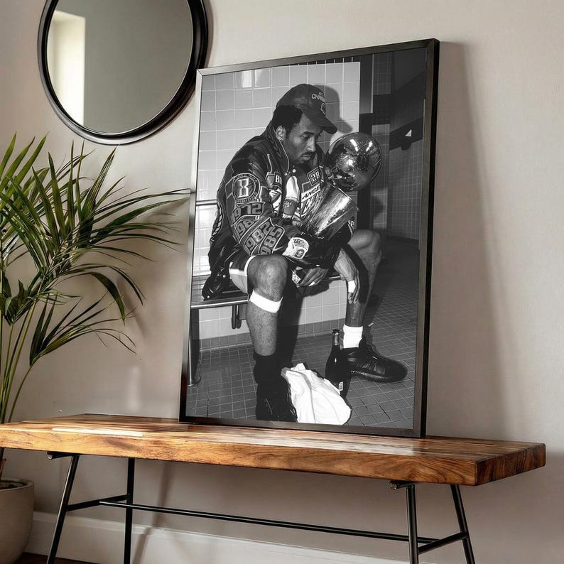 May include: Black and white framed photograph of a person seated, holding a trophy. The person is wearing a baseball cap and a jacket with patches. The photograph is displayed on a wooden bench with black metal legs, with a round mirror and a plant in the background.