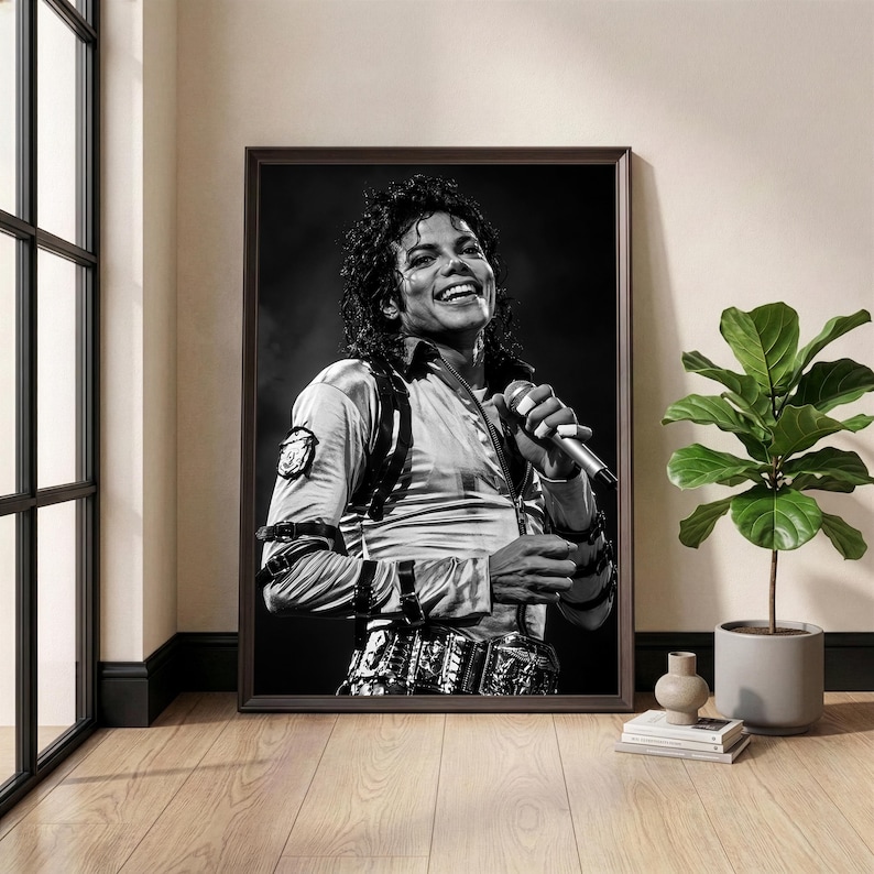 May include: Black and white framed print of a smiling person holding a microphone. The person is wearing a satin shirt and a decorative belt. The print is displayed on a wooden floor next to a potted plant.