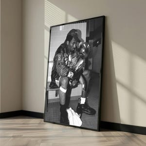 May include: Black and white framed photograph of a person sitting and holding a trophy. The person is wearing a jacket, shorts, and a baseball cap. The trophy is large and metallic. The photograph is leaning against a wall.
