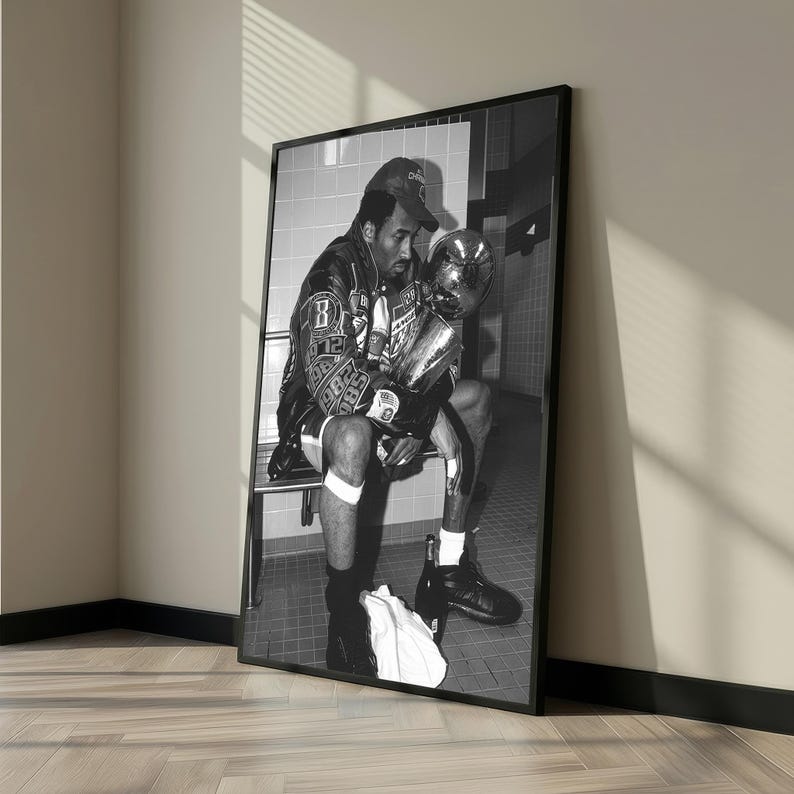 May include: Black and white framed photograph of a person sitting and holding a trophy. The person is wearing a jacket, shorts, and a baseball cap. The trophy is large and metallic. The photograph is leaning against a wall.