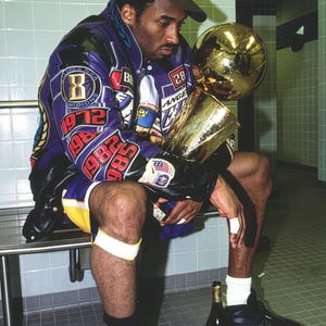 May include: A person wearing a purple and black jacket with patches and a championship hat, holding a gold trophy. The jacket has the number 8 and the years 1972, 1982, and 1985. Black shoes and a bottle of champagne are visible.