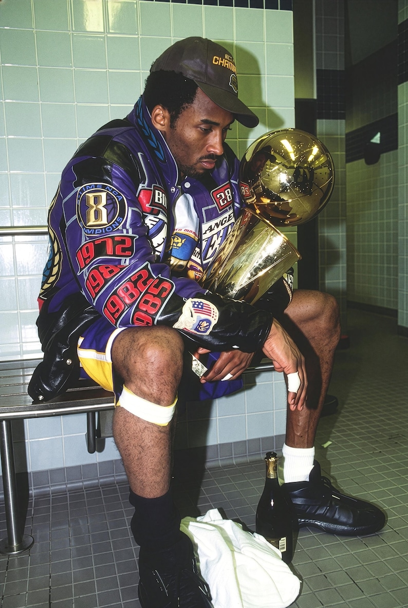 May include: A person wearing a purple and black jacket with patches and a championship hat, holding a gold trophy. The jacket has the number 8 and the years 1972, 1982, and 1985. Black shoes and a bottle of champagne are visible.