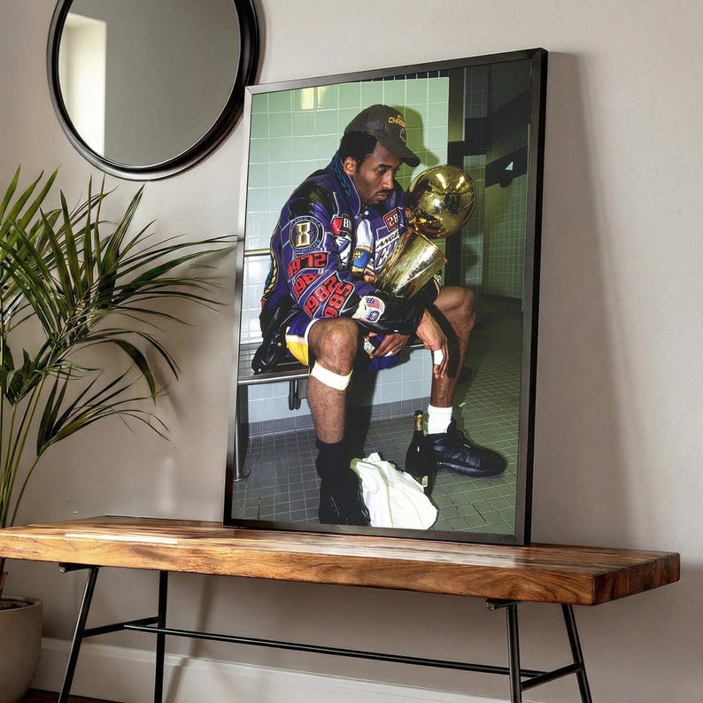 May include: Framed print featuring a seated figure holding a golden trophy. The figure wears a purple and gold jacket and a baseball cap. The image is displayed on a wooden bench with a black metal frame, with a round mirror and a plant in the background.
