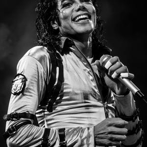 May include: Black and white image of a performer on stage, smiling and holding a microphone. The performer wears a silver jacket with black straps and a decorative belt. The image is a medium shot, capturing the performer's upper body.