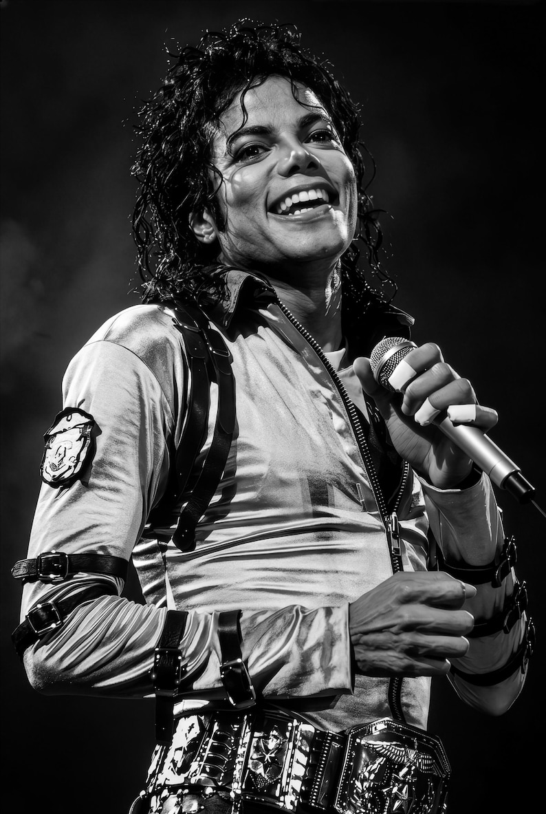 May include: Black and white image of a performer on stage, smiling and holding a microphone. The performer wears a silver jacket with black straps and a decorative belt. The image is a medium shot, capturing the performer's upper body.