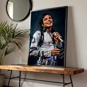 May include: Framed portrait of Michael Jackson, smiling and holding a microphone. He wears a silver jacket with black accents and a decorative belt. The portrait is displayed on a wooden bench with black metal legs, near a plant and a round mirror.