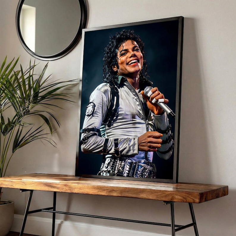 May include: Framed portrait of Michael Jackson, smiling and holding a microphone. He wears a silver jacket with black accents and a decorative belt. The portrait is displayed on a wooden bench with black metal legs, near a plant and a round mirror.