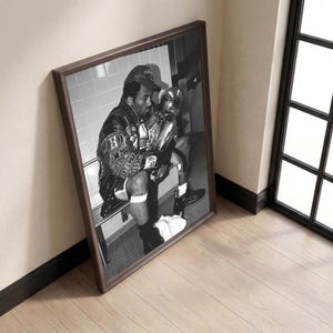 May include: Black and white framed print of a man in a leather jacket and baseball cap, holding a trophy. The image is a portrait of a man sitting on a bench in a locker room setting.