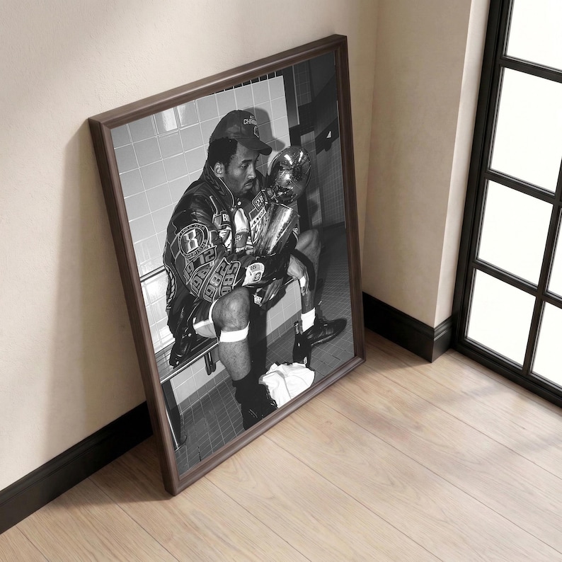 May include: Black and white framed print of a man in a leather jacket and baseball cap, holding a trophy. The image is a portrait of a man sitting on a bench in a locker room setting.
