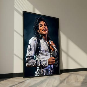 May include: Framed portrait of Michael Jackson smiling and holding a microphone. He is wearing a silver jacket with black accents and a decorative belt. The image is in a black frame, leaning against a white wall.