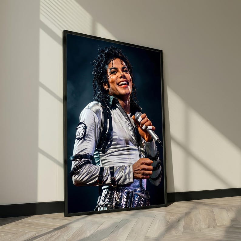May include: Framed portrait of Michael Jackson smiling and holding a microphone. He is wearing a silver jacket with black accents and a decorative belt. The image is in a black frame, leaning against a white wall.