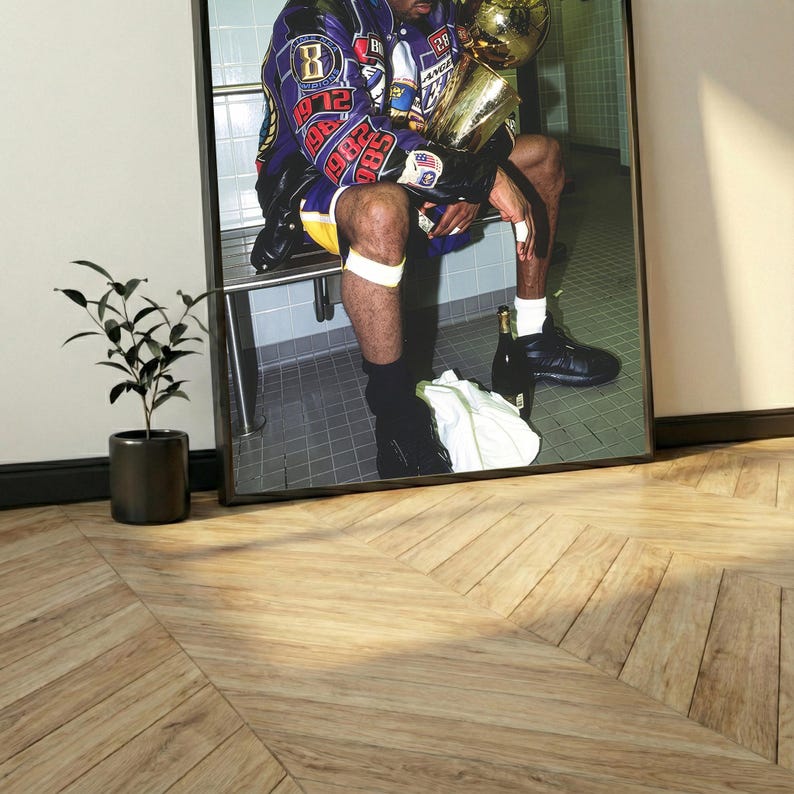 May include: Framed photograph of a person in a purple and gold jacket holding a gold trophy. The person is seated, with a white towel and a bottle on the floor. The image is in a black frame, set against a white wall and a wooden floor.