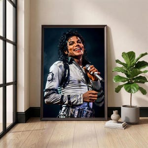 May include: Framed print of Michael Jackson performing on stage, smiling and holding a microphone. He wears a silver jacket with black accents and a decorative belt. The image is in a dark brown frame, displayed in a room with a window and a potted plant.
