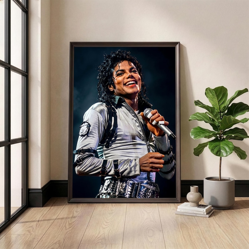 May include: Framed print of Michael Jackson performing on stage, smiling and holding a microphone. He wears a silver jacket with black accents and a decorative belt. The image is in a dark brown frame, displayed in a room with a window and a potted plant.